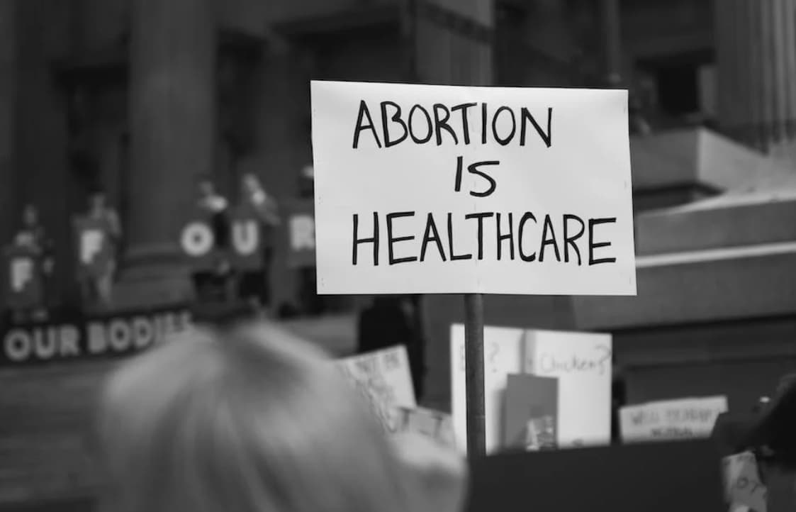 A black and white photo of a protest scene focusing on a sign that reads "ABORTION IS HEALTHCARE"